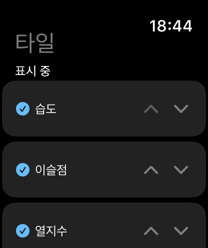‘타일’ 제목과 ‘표시 중’ 아래에 습도, 이슬점, 열지수가 각각 체크되어 있고 위아래 꺾쇠 모양 화살표가 있는 워치의 타일 화면.